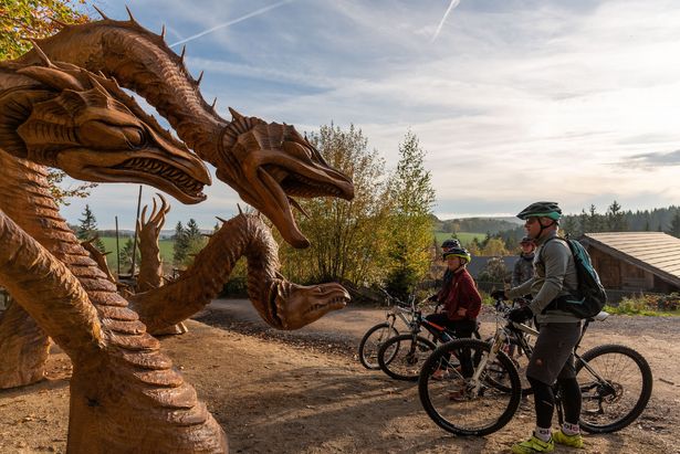 Erzgebirge Blockline Holzfiguren Eine Familie steht mit ihren Rädern am Rande der Blockline-Tour. Sie schauen auf Drachenfiguren aus Holz.