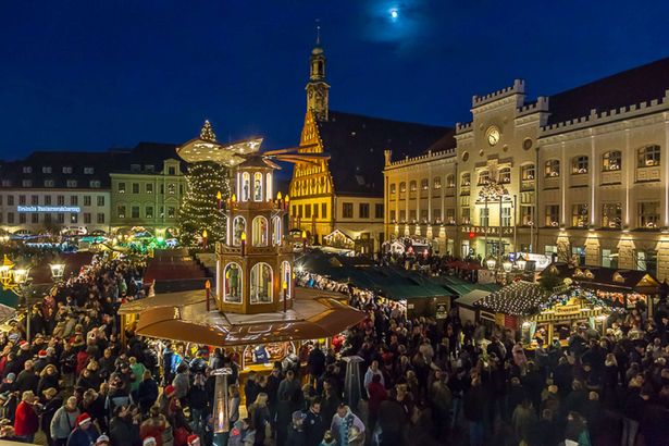 A market square is decorated for Christmas. In its center is a large Christmas pyramid. Various stalls and stalls line the square. There are lots of people on the go.